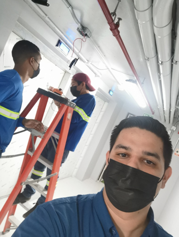 Three workers in blue uniforms on a ladder in a bright industrial hallway, one taking a selfie.