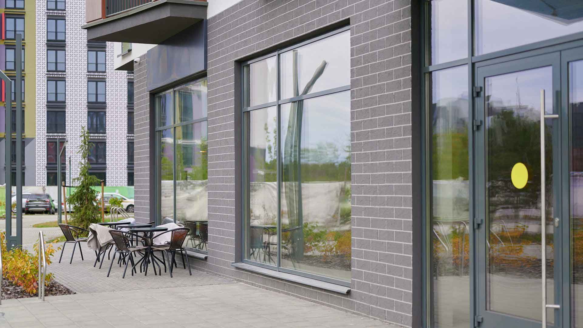 Exterior of a building with large windows and a seating area outside. Gray brick facade, glass doors, and tables/chairs. Exterior of a building with large windows and a seating area outside. Gray brick facade, glass doors, and tables/chairs.