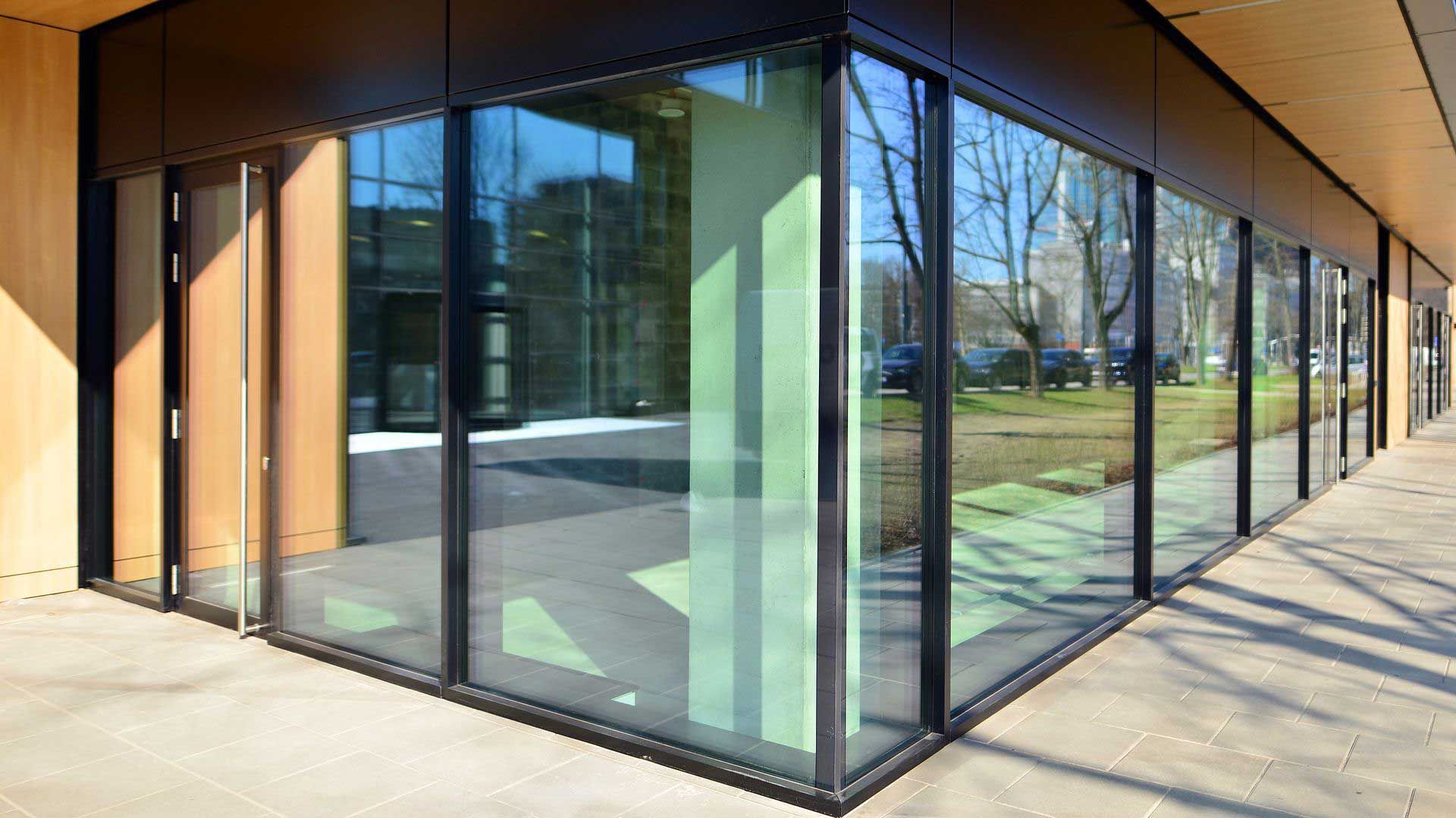 Glass-fronted building with black frames and empty interior reflecting surroundings. Plants in foreground, paved walkway. Glass-fronted building with black frames and empty interior reflecting surroundings. Plants in foreground, paved walkway.