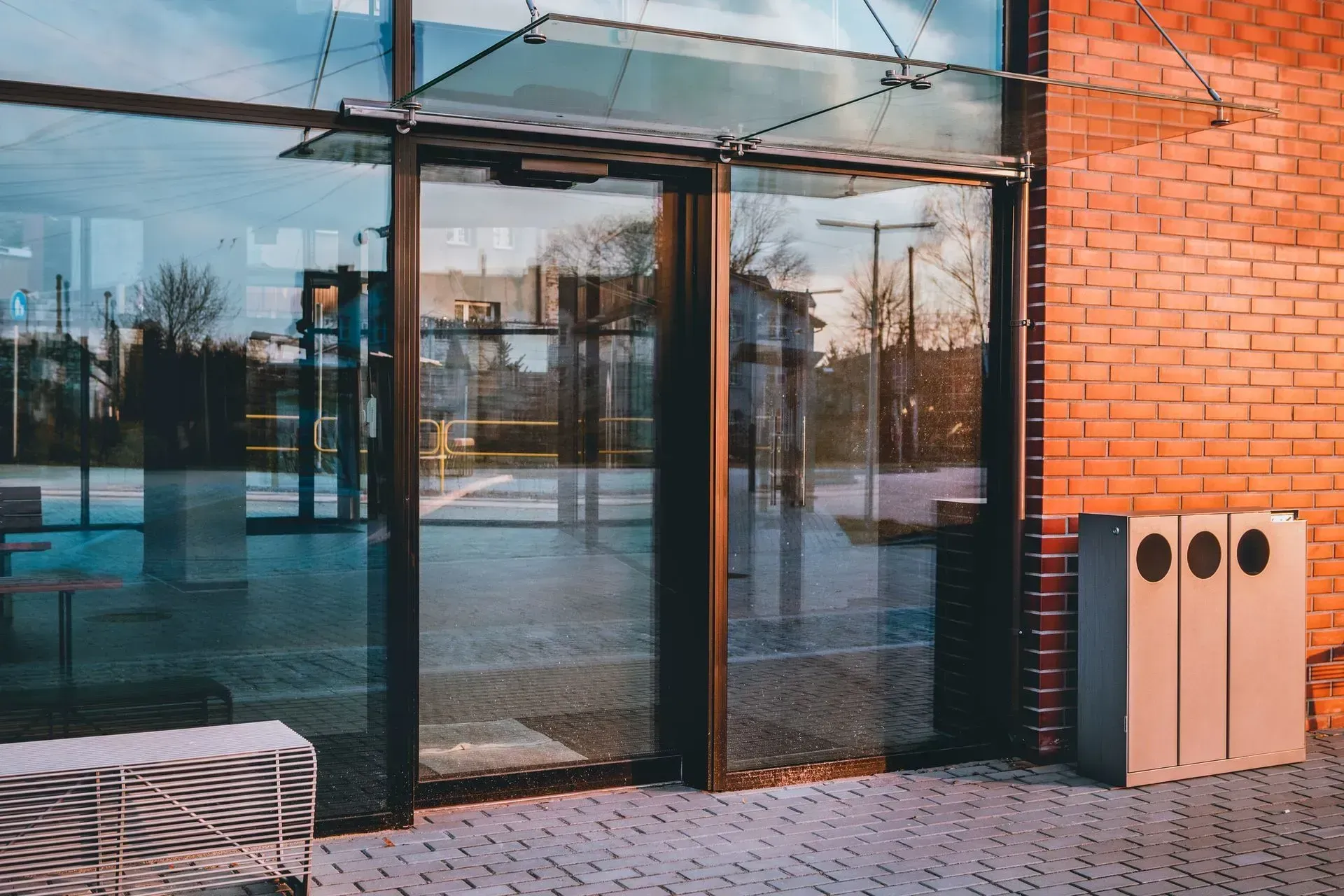 Glass doors and brick wall, exterior building view. Three recycling bins. Paved walkway. Sunlight.