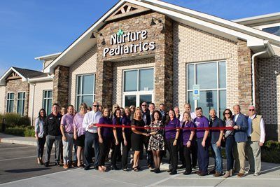 A group of people holding a red ribbon during a grand opening ceremony in front of the Nurture Pediatrics building.