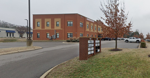 A two-story red brick office building with a parking lot and a directory sign on a cloudy day.