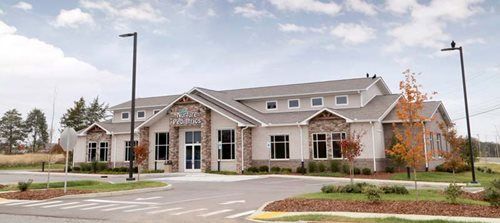 A single-story, light-colored commercial building with stone accents, gray roofing, and a paved parking lot in front.