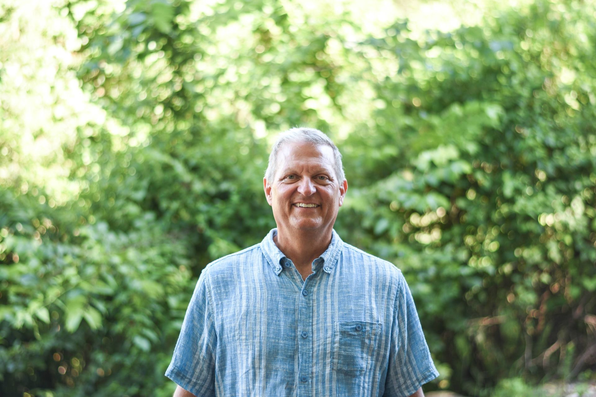 Smiling man in a blue striped shirt standing outdoors in a green garden.