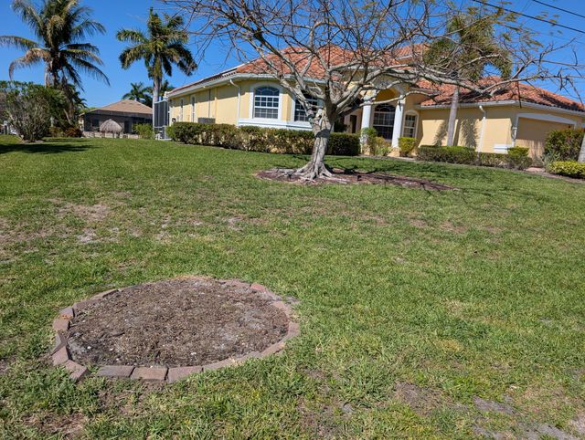 Single-story yellow house with red tile roof and palm trees beside a grassy front yard