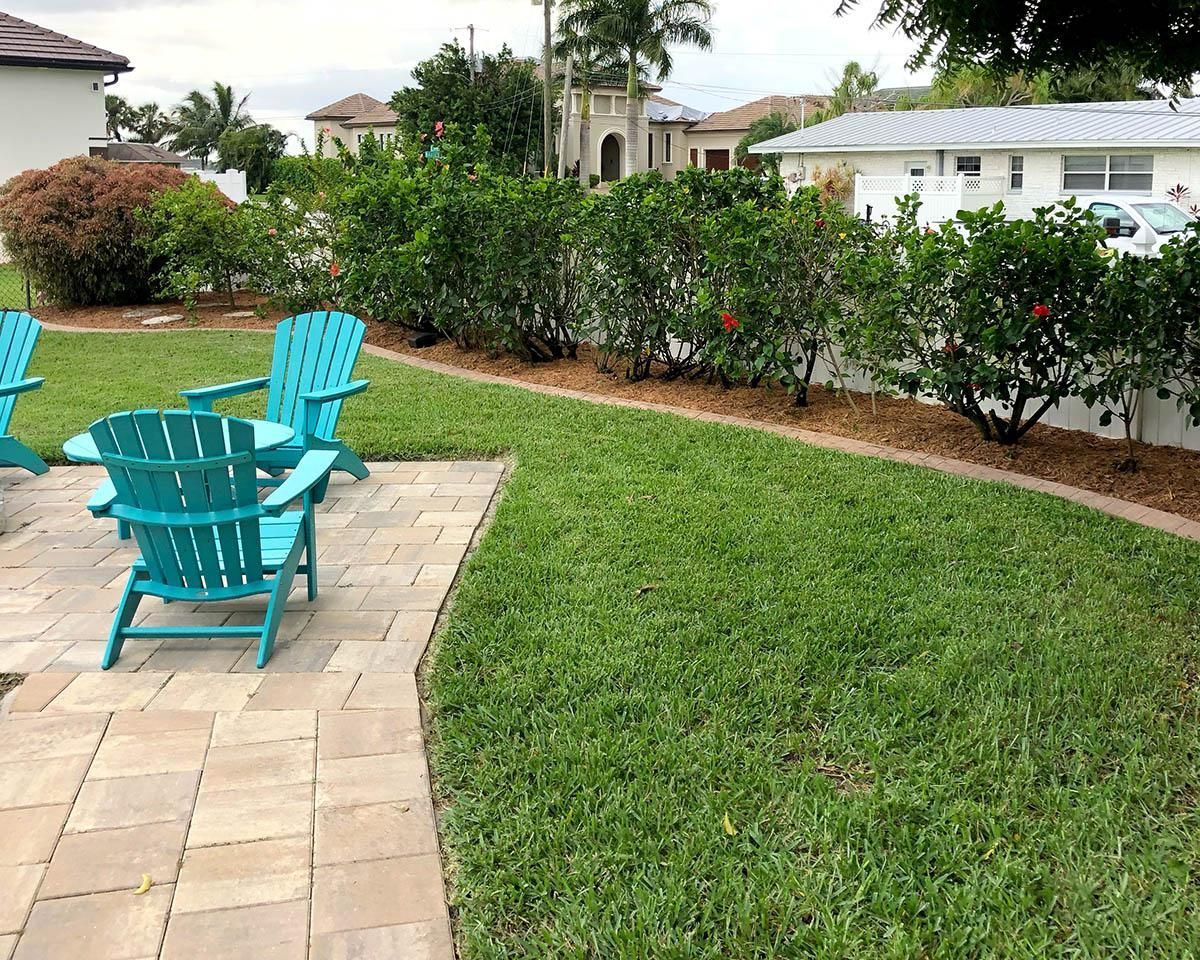 Two bright teal Adirondack chairs sit on a light stone patio next to a lush green lawn and a manicured hedge.