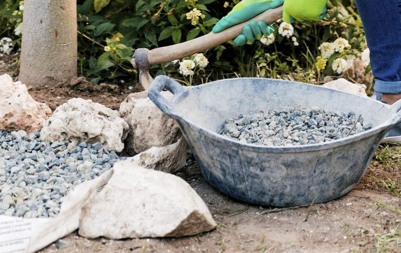 A gloved hand uses a hoe to move small grey stones near a basin full of gravel in a garden setting.