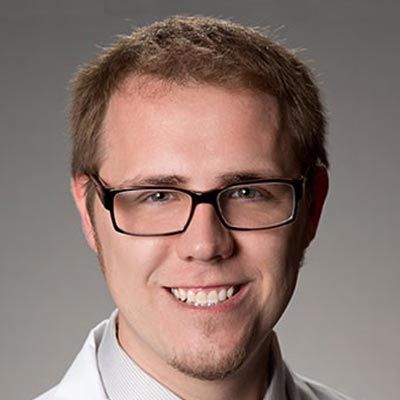 Man with glasses smiles, wearing a white coat and light shirt; soft background.