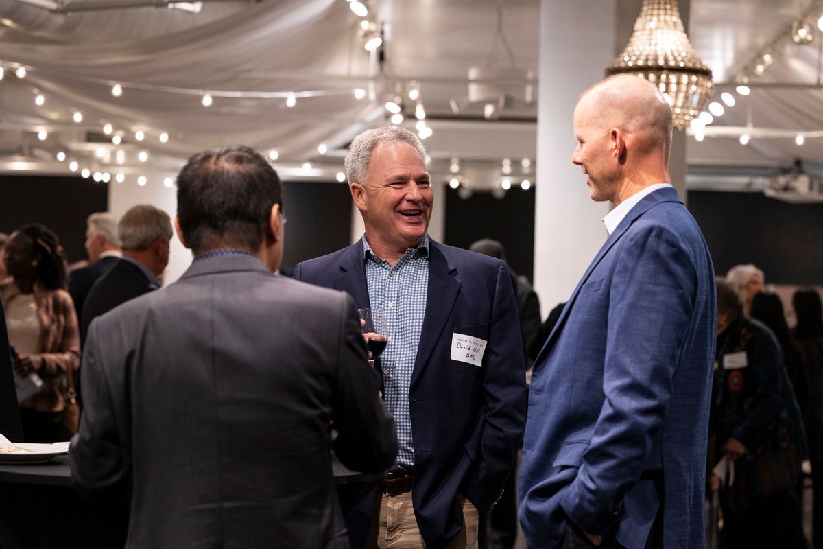 Three men in suits chatting at a well-lit event. One man smiles, holding a drink. Other people are in the background.