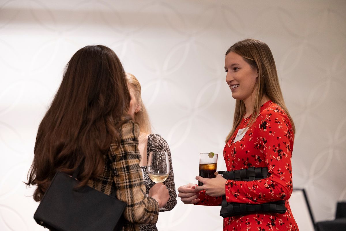 Three women chatting at an event. One in a red dress holds a drink, the others face her.