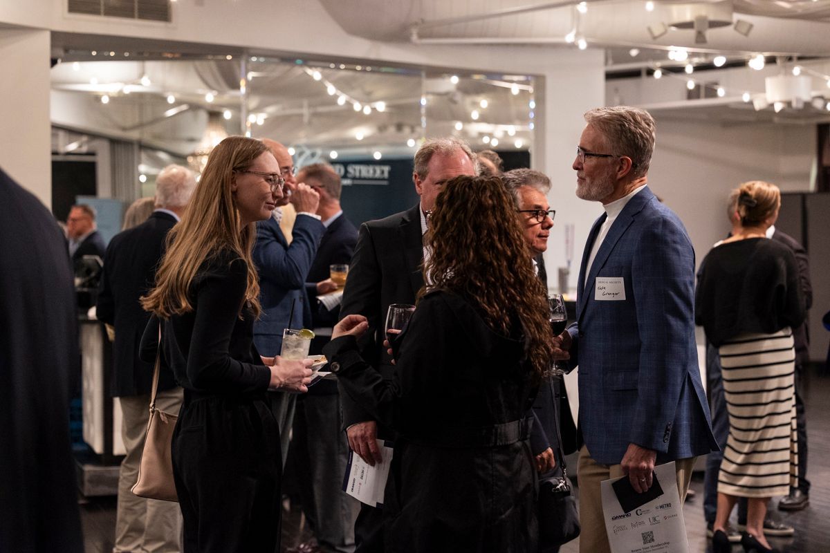 People mingling at an event, holding drinks, in a well-lit room with a mirror and lights.