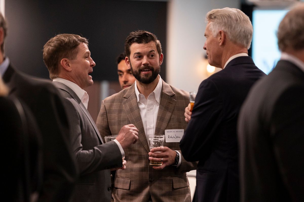 Men in suits at a social gathering, engaged in conversation, holding drinks.