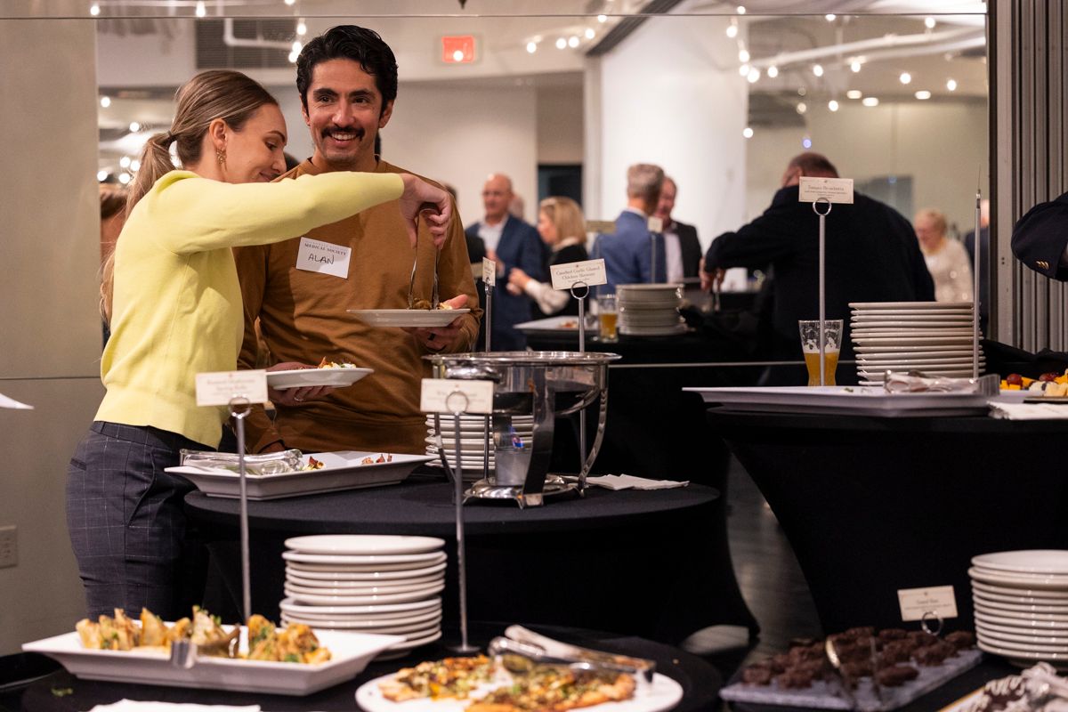 People at an event, selecting food from buffet tables. Woman offers man a sample.