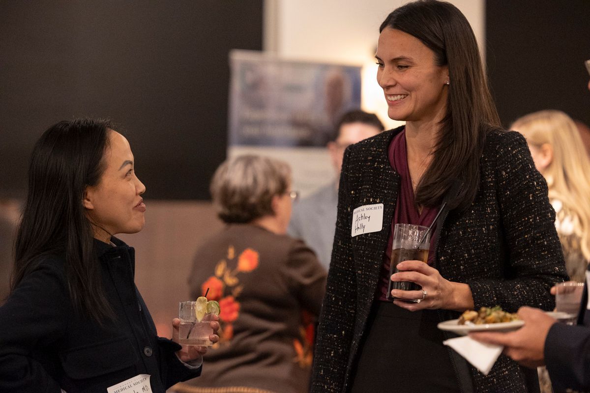 Two women at an event, one holding a drink and plate of food, smiling and talking.