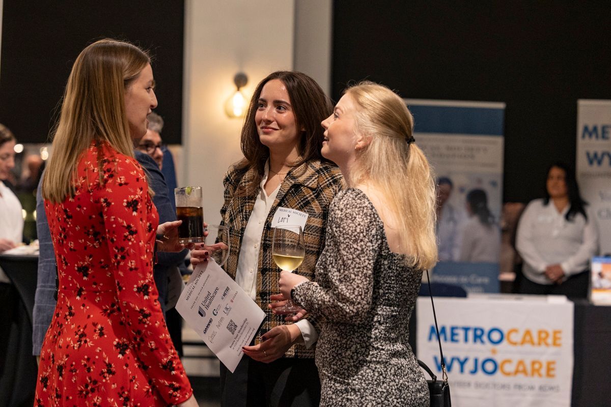Three women in a social setting; one in red dress, others in patterned tops, holding drinks, talking.