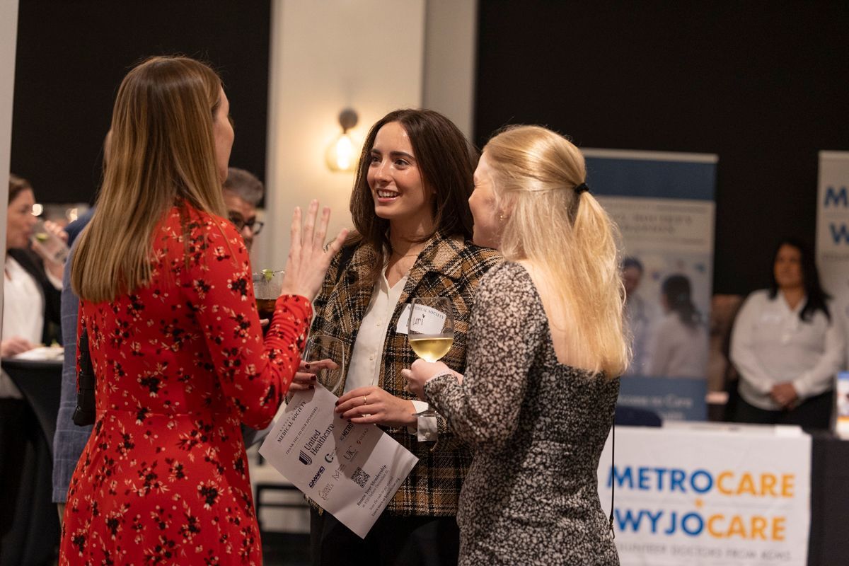 Three women conversing at an event, one holding a drink and a paper, standing near display banners.