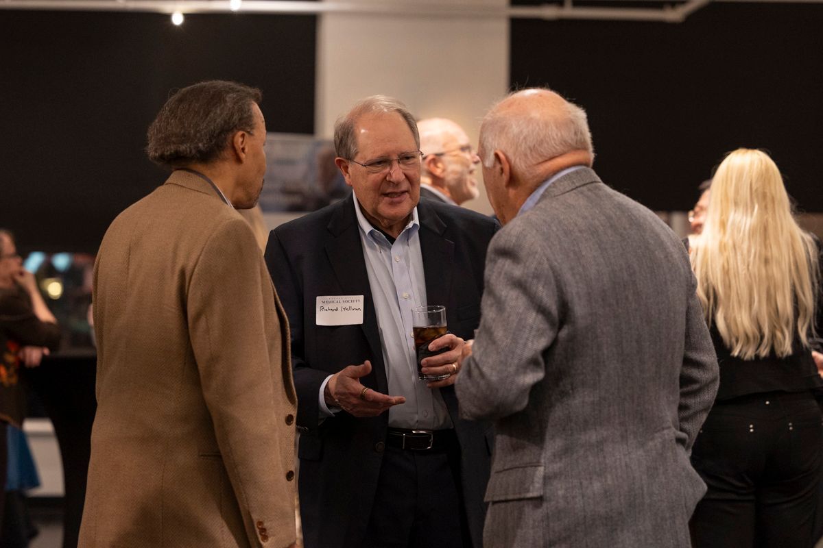 Three men in suits converse at an event, one holding a drink.