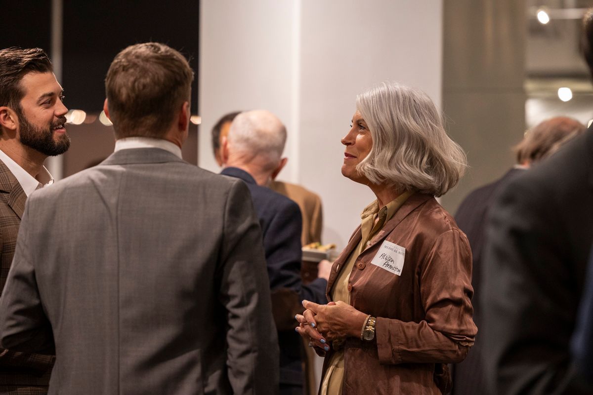 People at an event, talking and smiling; woman in brown jacket, other people in suits, in a room.