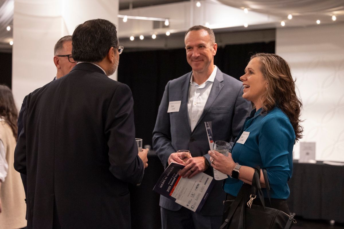 Group of people networking at an event. They are standing and talking, some holding drinks or papers.