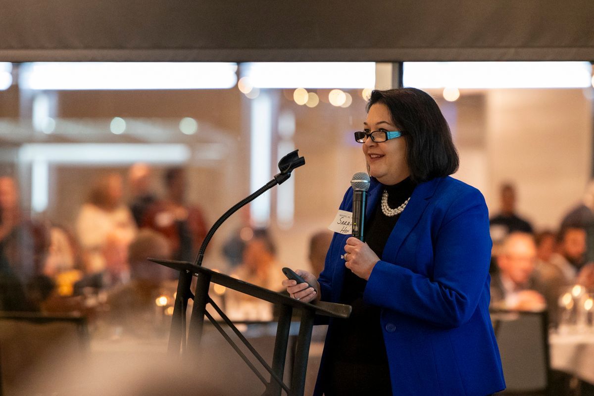 Woman speaking at a podium, wearing a blue blazer, pearl necklace, and glasses. Crowd in background.