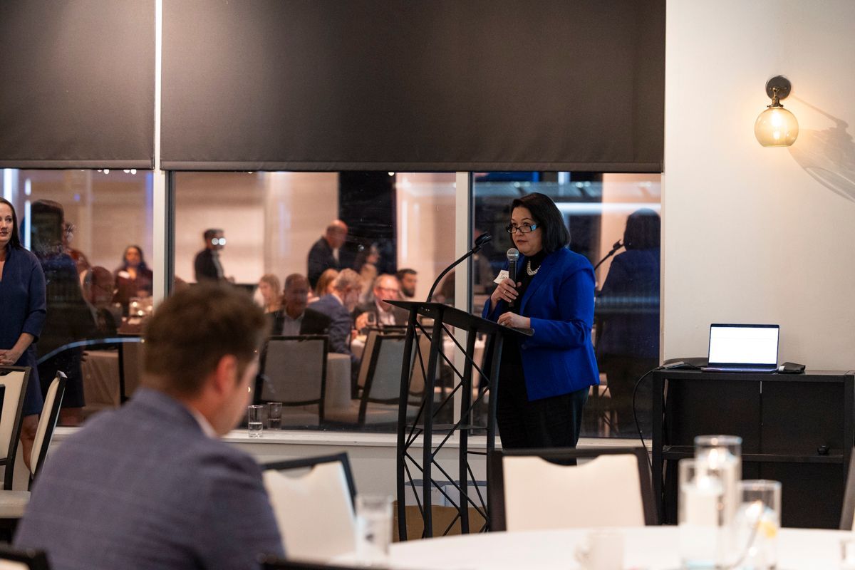 Woman speaking at a podium, addressing a seated audience in a venue with large windows.