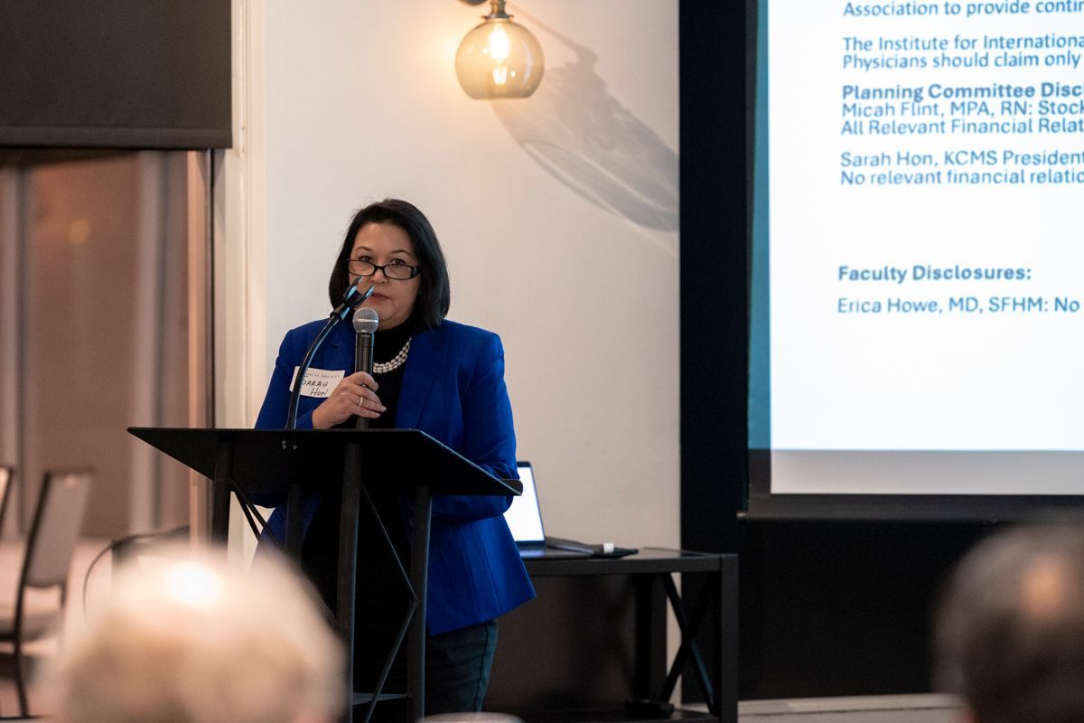 Woman in blue blazer speaks at a podium; presentation screen in background.