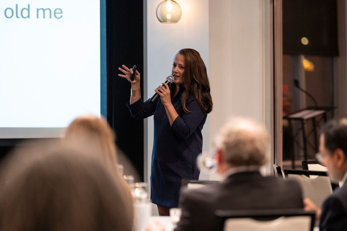 Woman speaking into a microphone, gesturing at a screen in a conference setting.