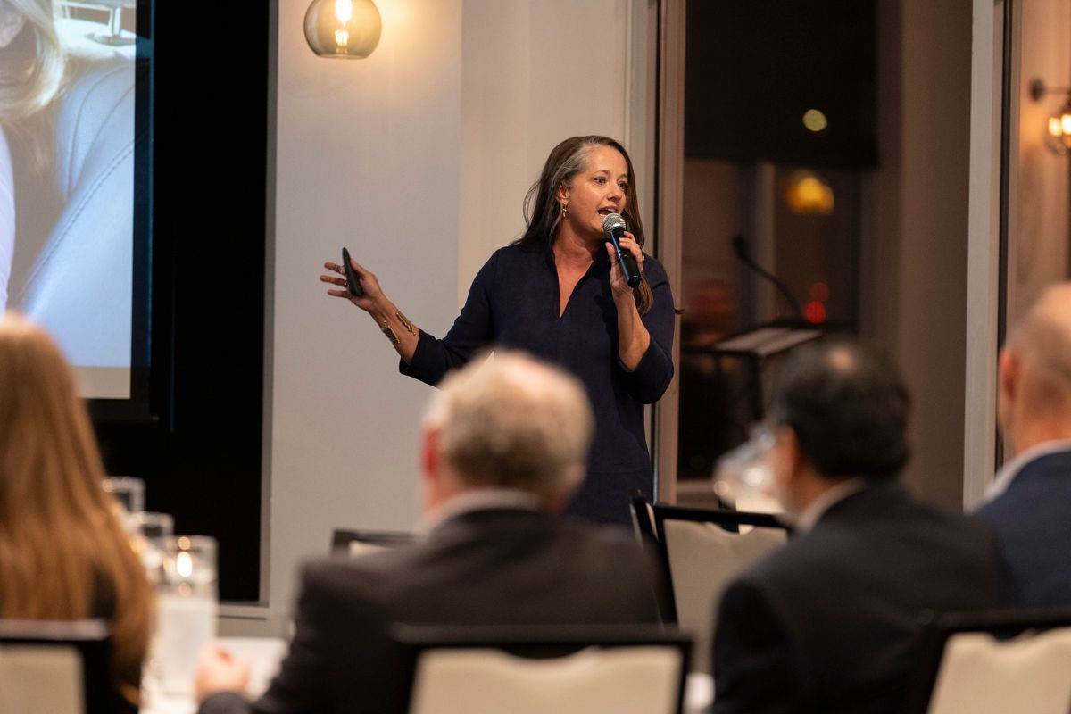 Woman speaking at a presentation, gesturing with one hand, holding a microphone. Audience seated at tables.