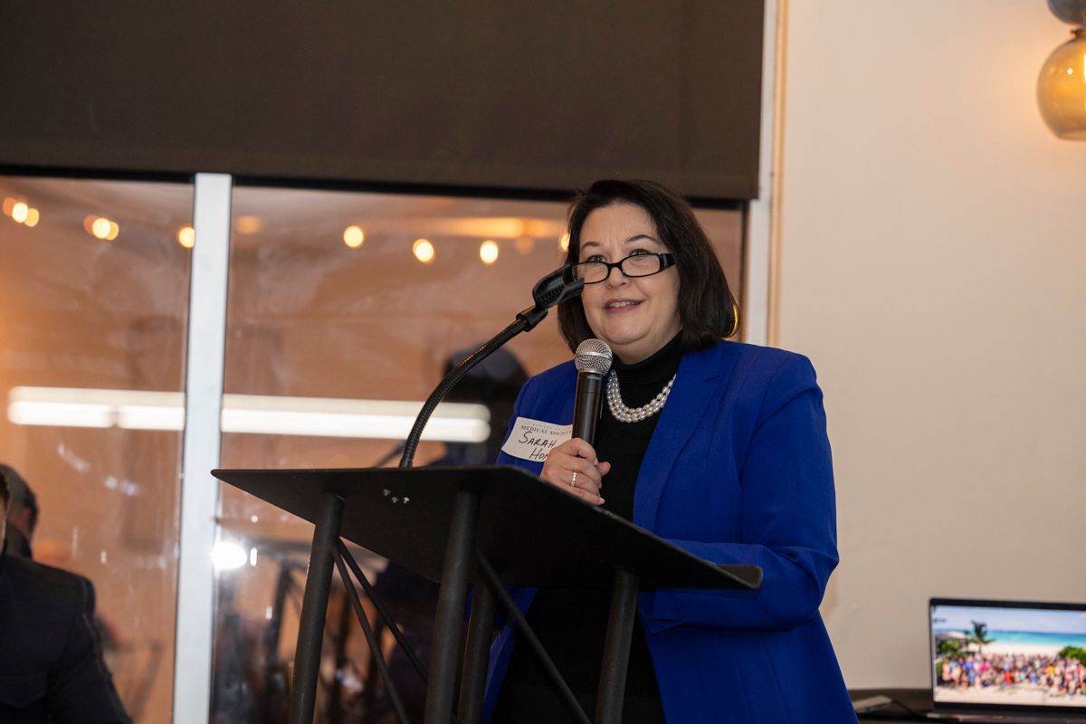 Woman in blue blazer speaks at a podium; interior setting with window and laptop.