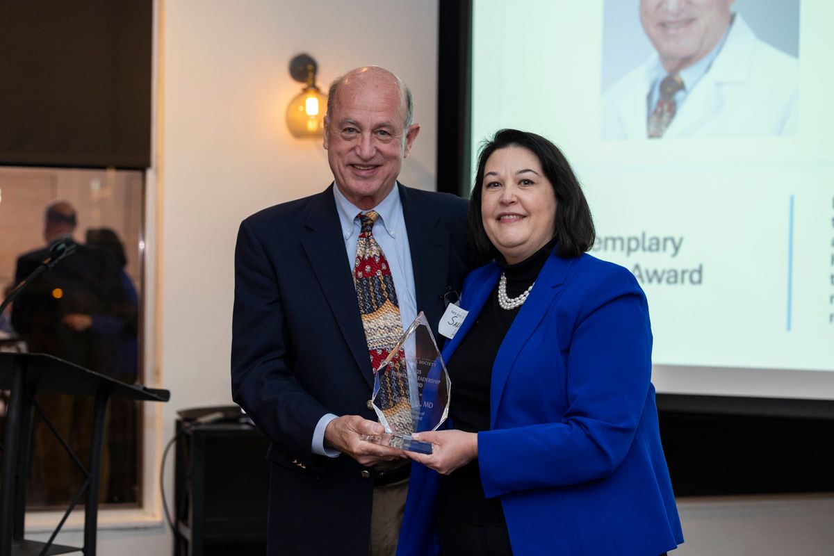 Man in suit presents an award to a woman in a blue jacket; both smiling near a screen.
