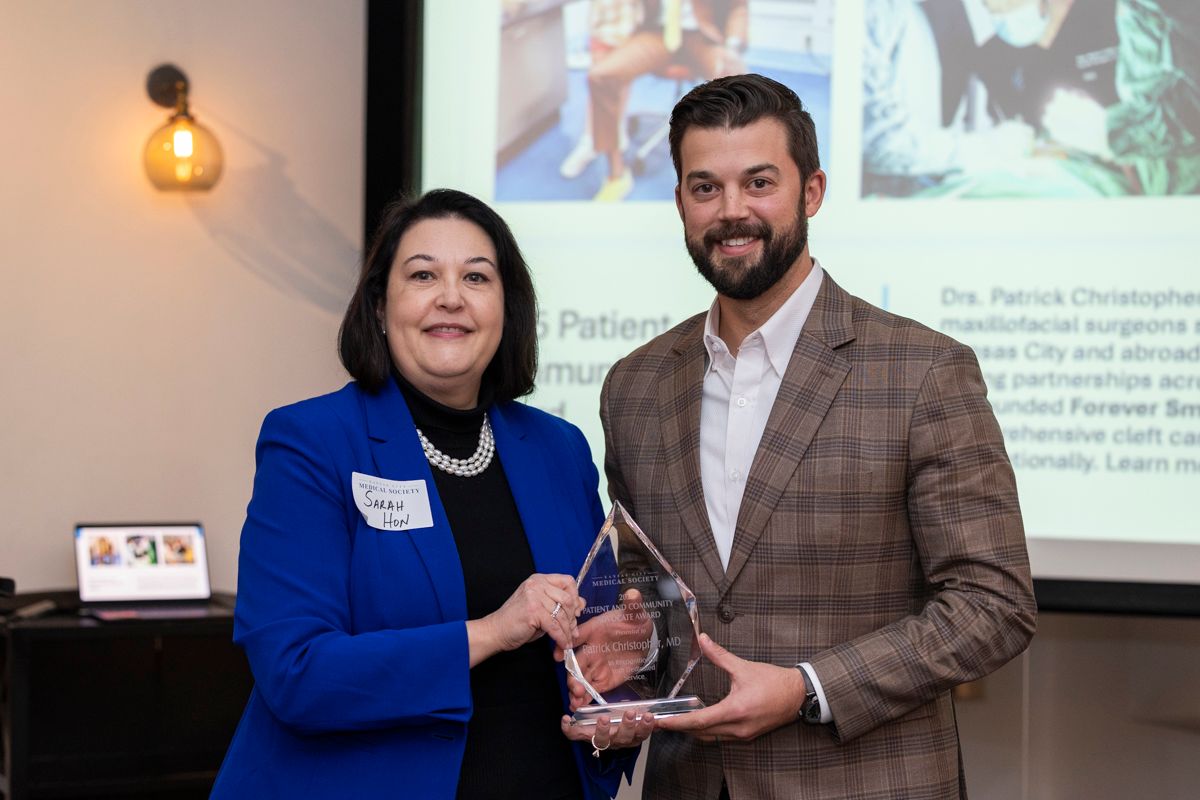 Woman in blue jacket presents award to man in a blazer. Blurred background with a screen.