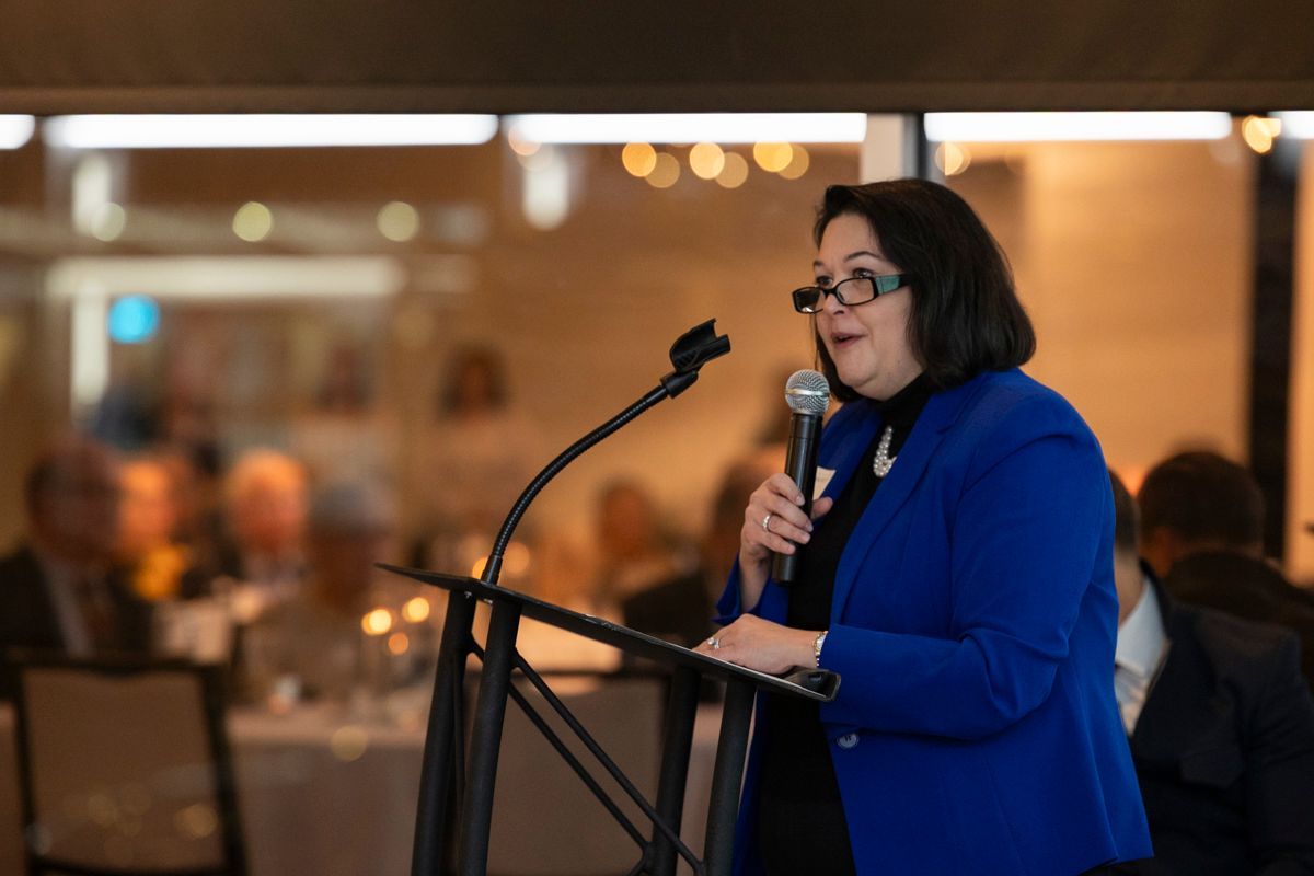 Woman in blue blazer speaks at a podium, holding a microphone. Inside a well-lit event.