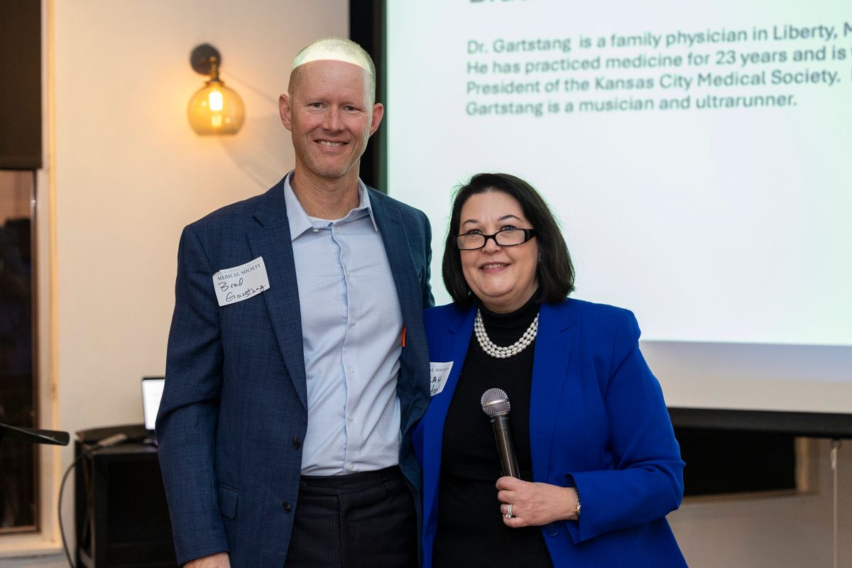 Two people standing, smiling, with a presentation screen in the background. Man in blazer; woman holding a microphone.