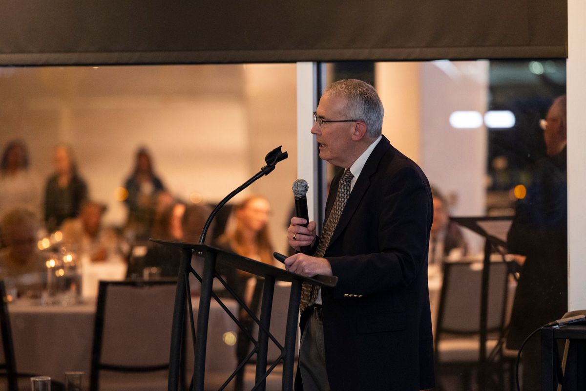 Man speaking at a podium with a microphone, indoors, with a seated audience in the background.