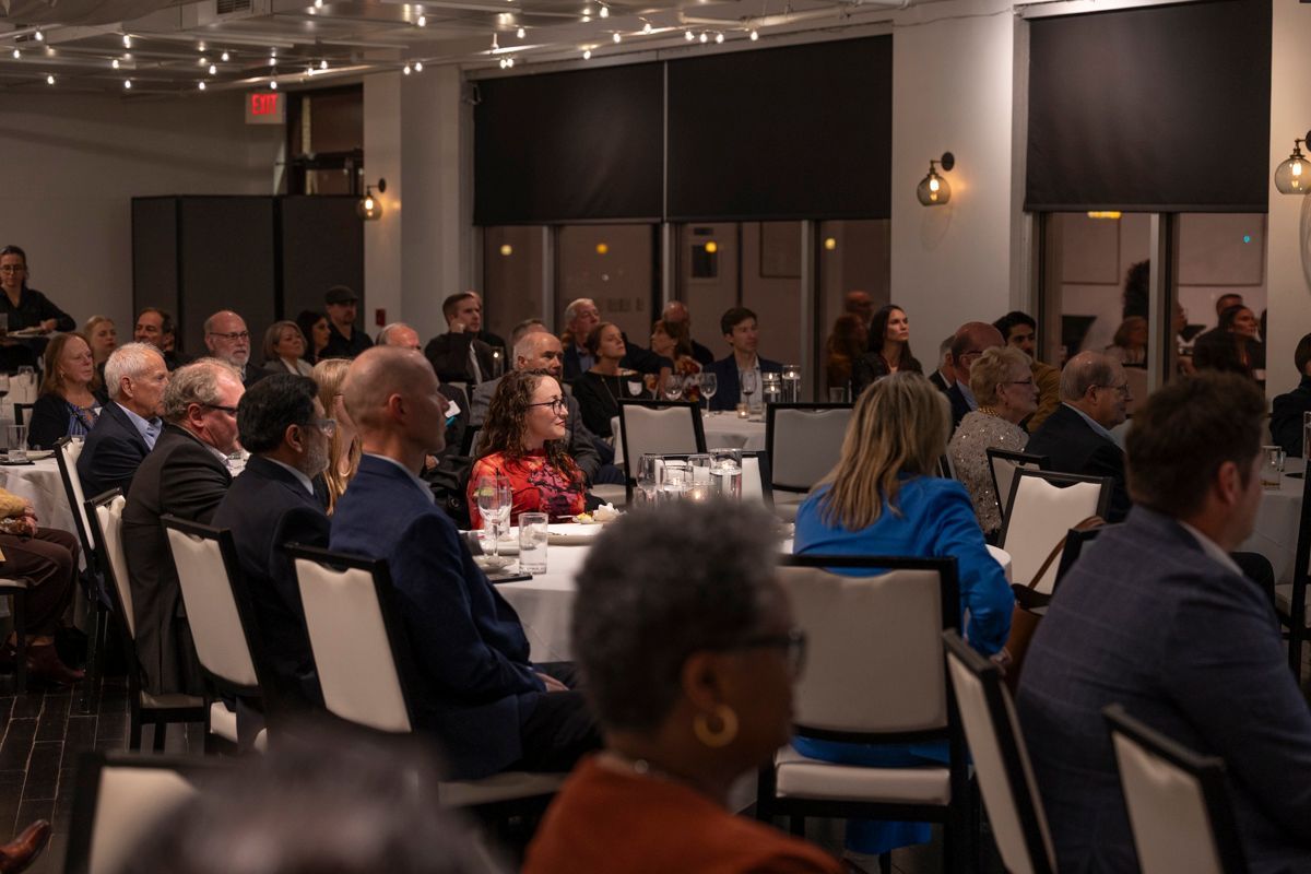 Audience at an event seated at round tables. Dim lighting, some people looking forward.