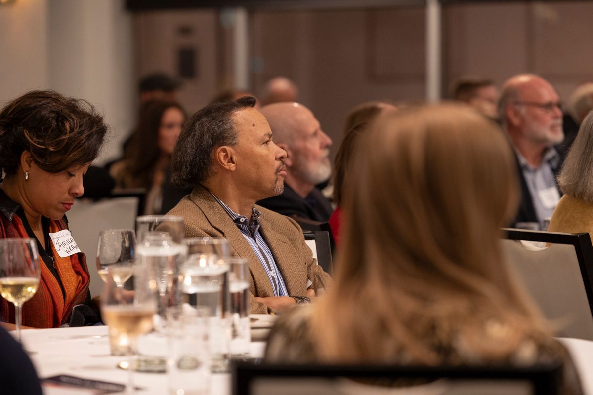 People seated at tables, listening at a conference. Several have glasses of wine and water.