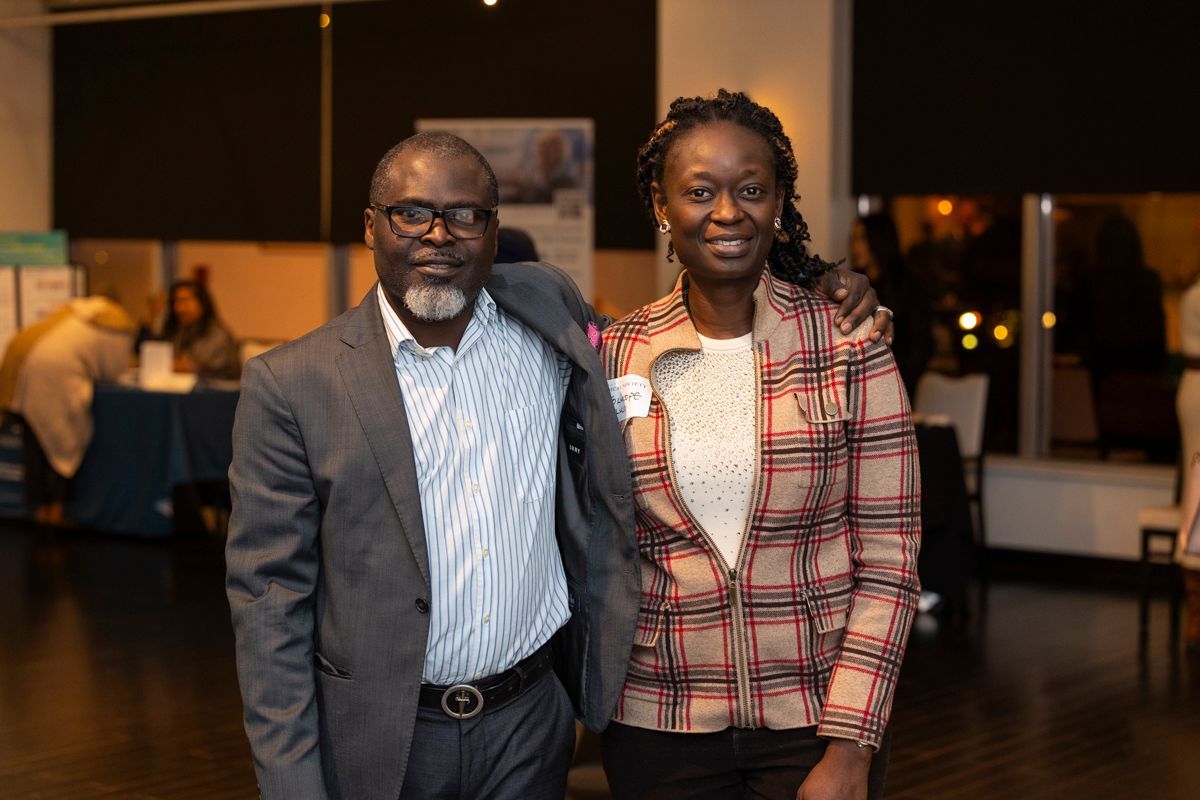 Man in suit and woman in plaid jacket, smiling, arm in arm in an event space.