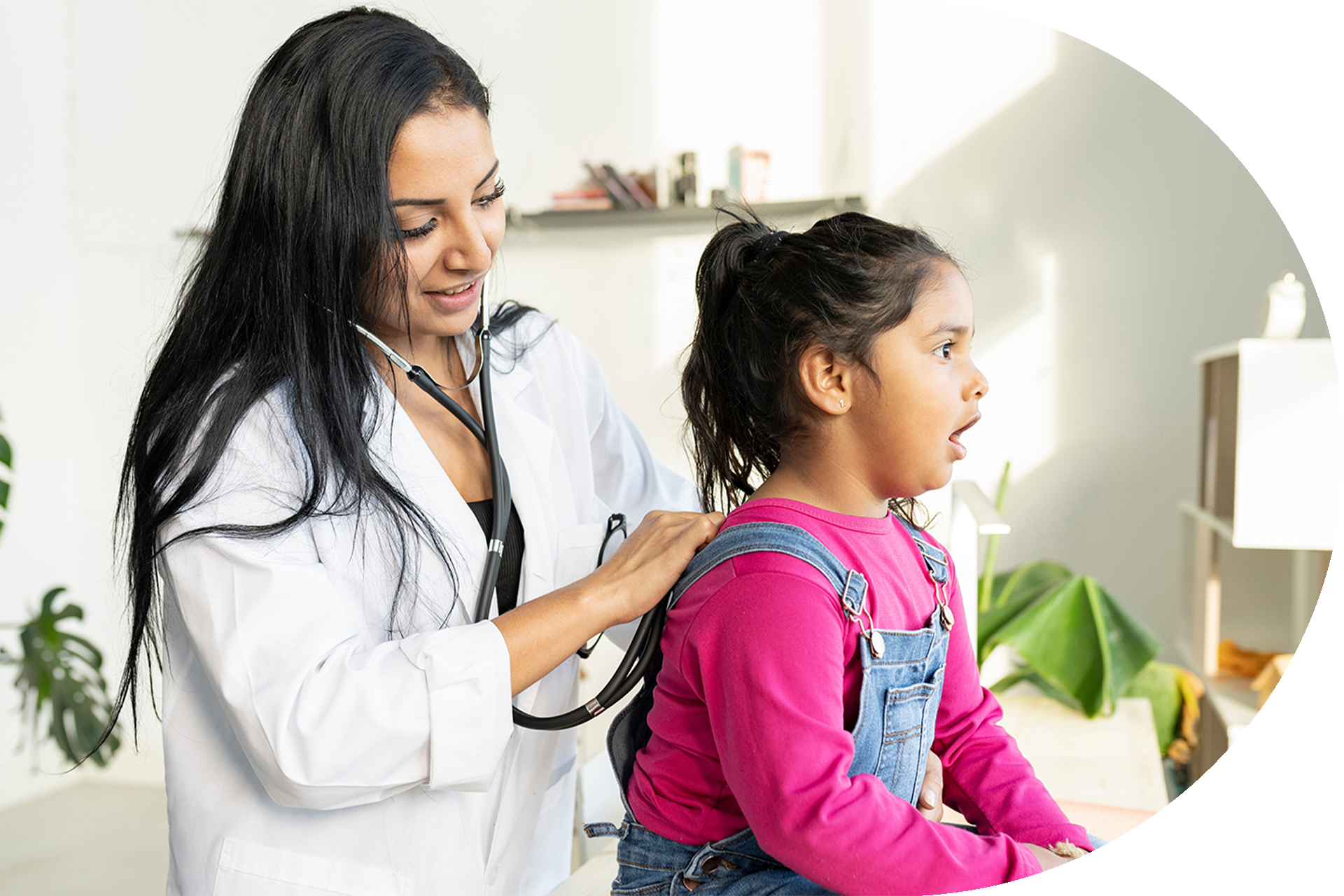 Doctor examining a child's back with a stethoscope in a bright room. The child looks up with their mouth open.