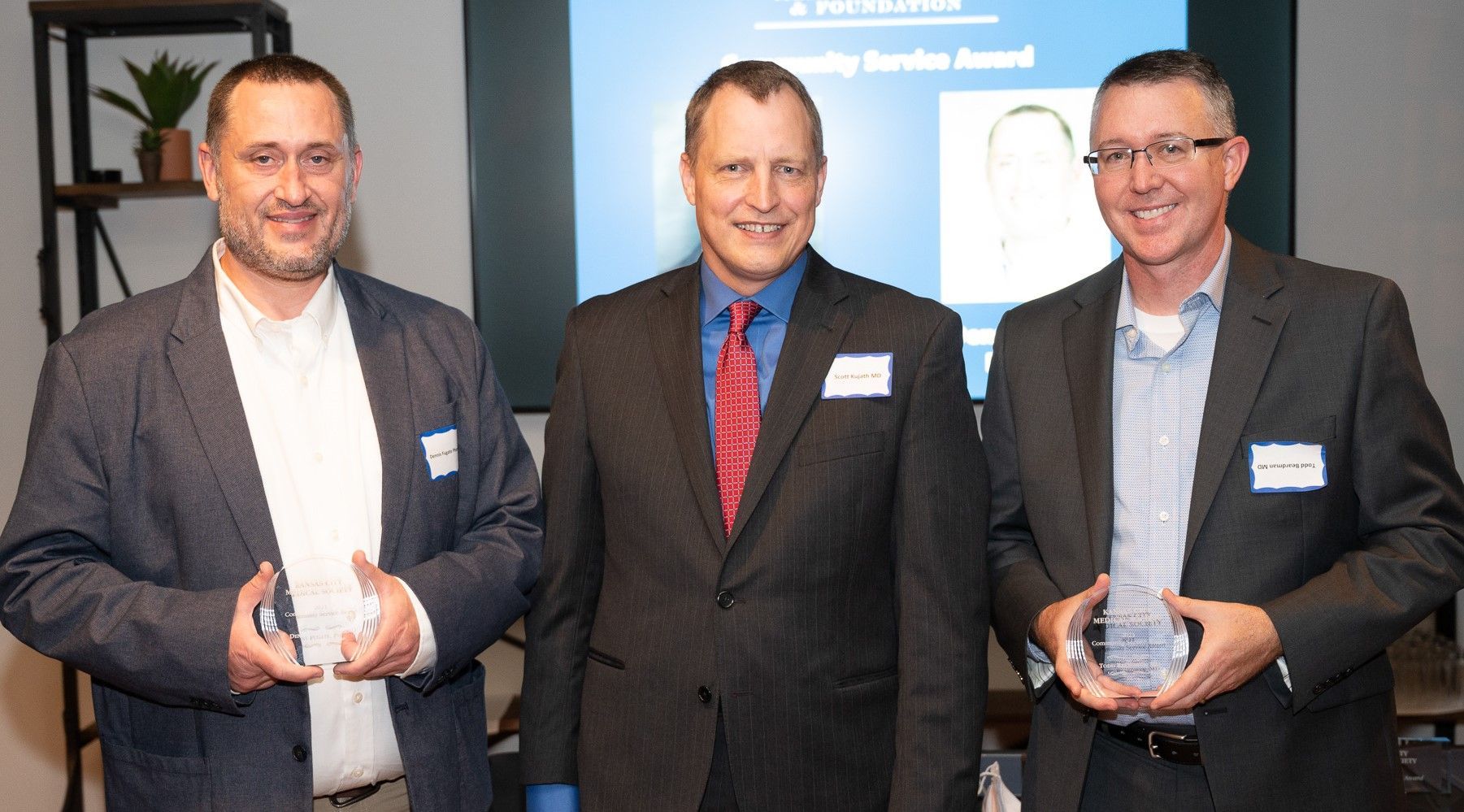 Three men in suits holding awards, smiling in front of a screen.