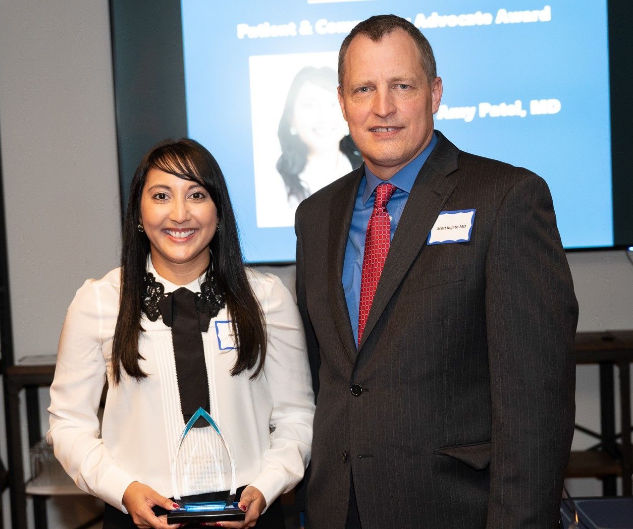Woman holding award, smiles alongside suited man at a ceremony; blue background with text.