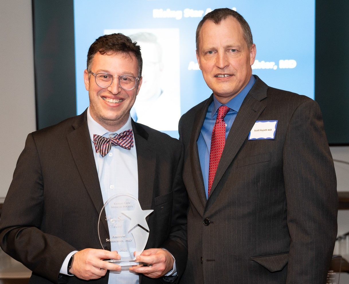 Two men in suits. One holds an award, both smiling. Blue backdrop with text.