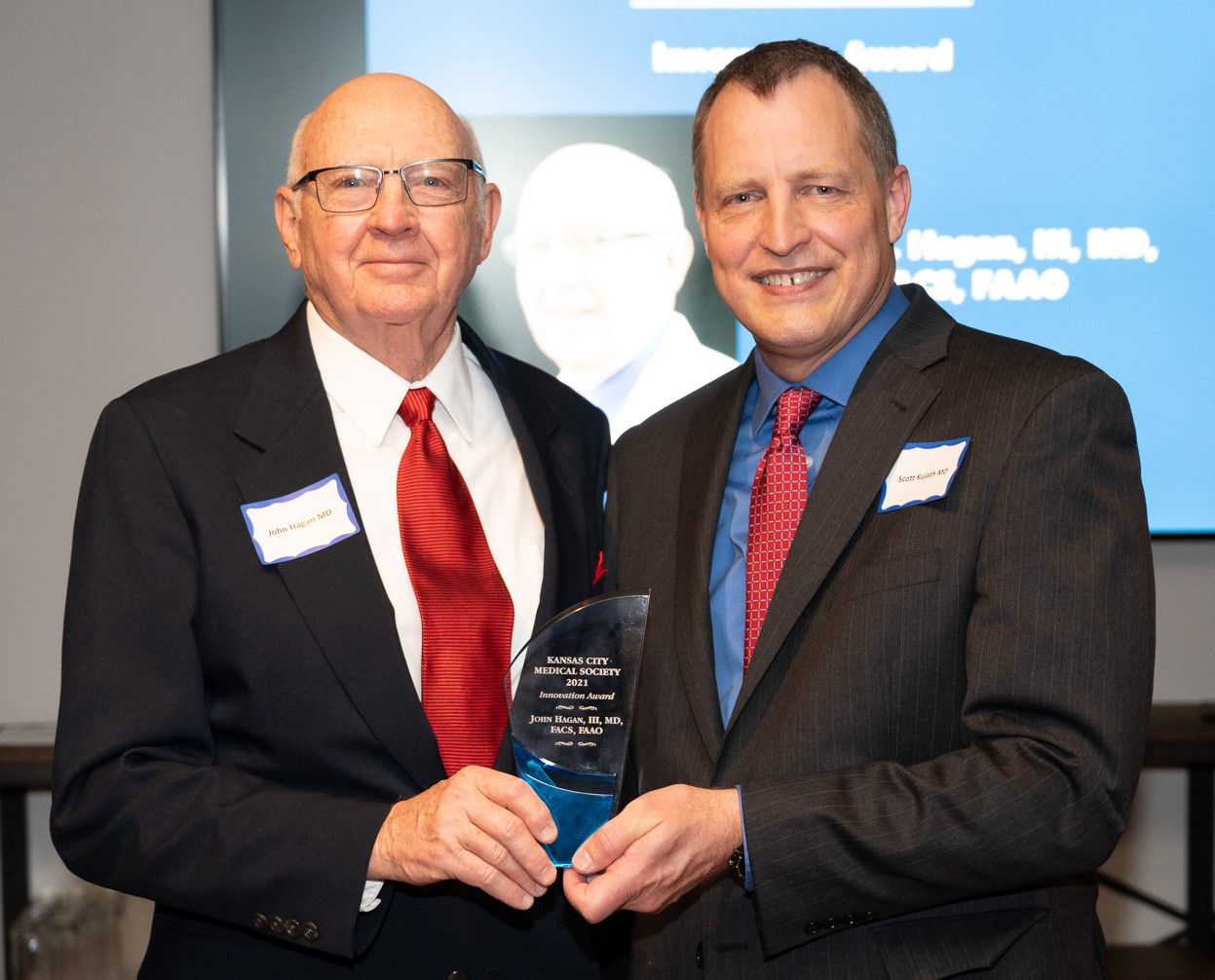 Two men in suits holding an award; a man on the right smiles.