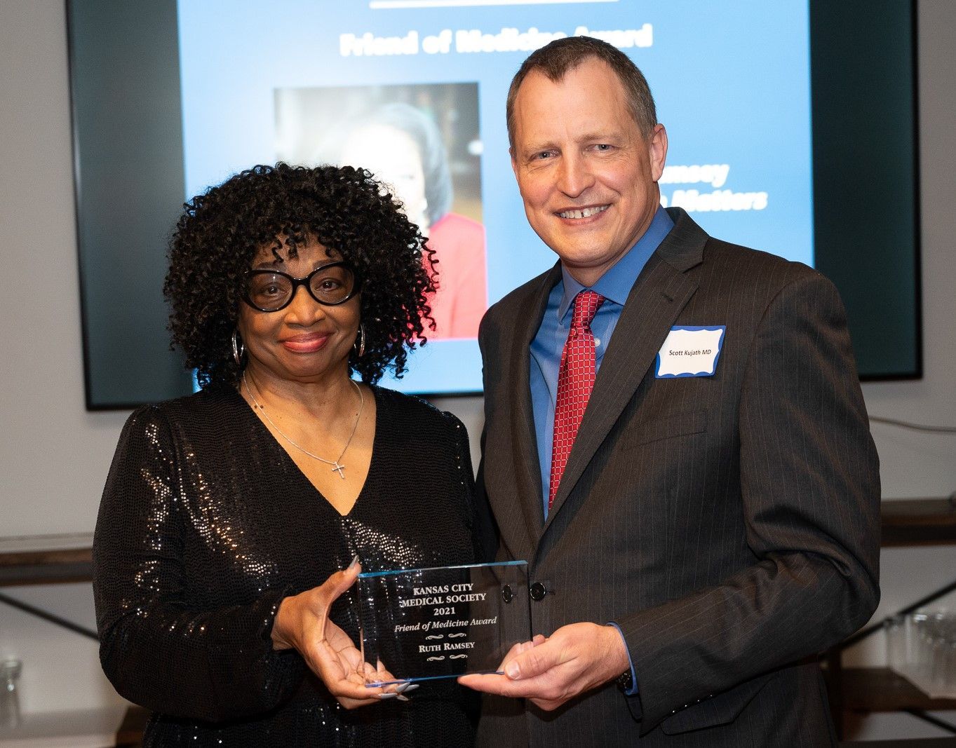 Woman in sequin top and man in suit holding an award. They are smiling at a ceremony.