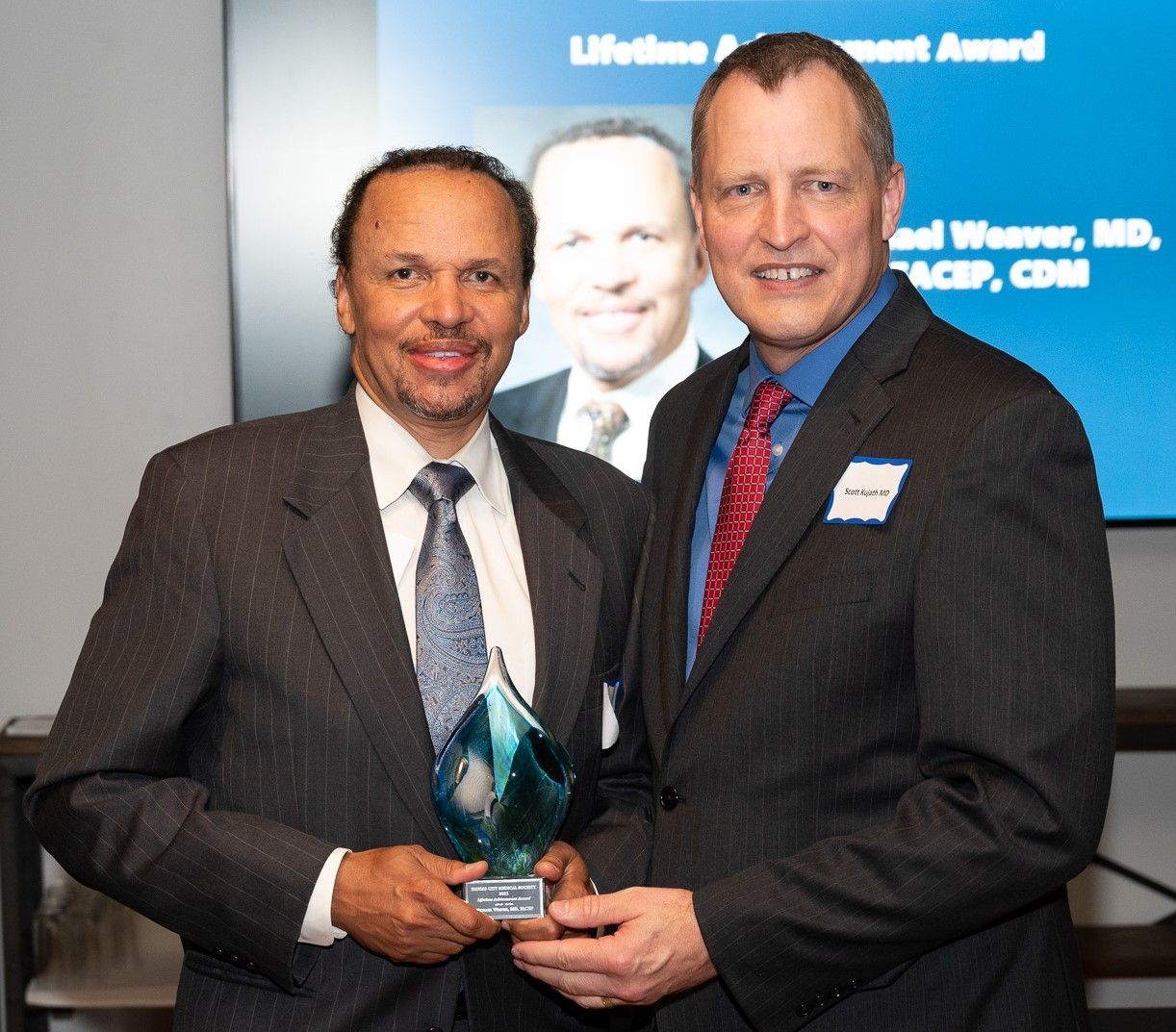Two men in suits holding an award; one smiling, a photo on screen.