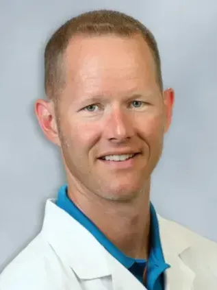 Man in a white coat smiling, light skin, short brown hair, blue polo.