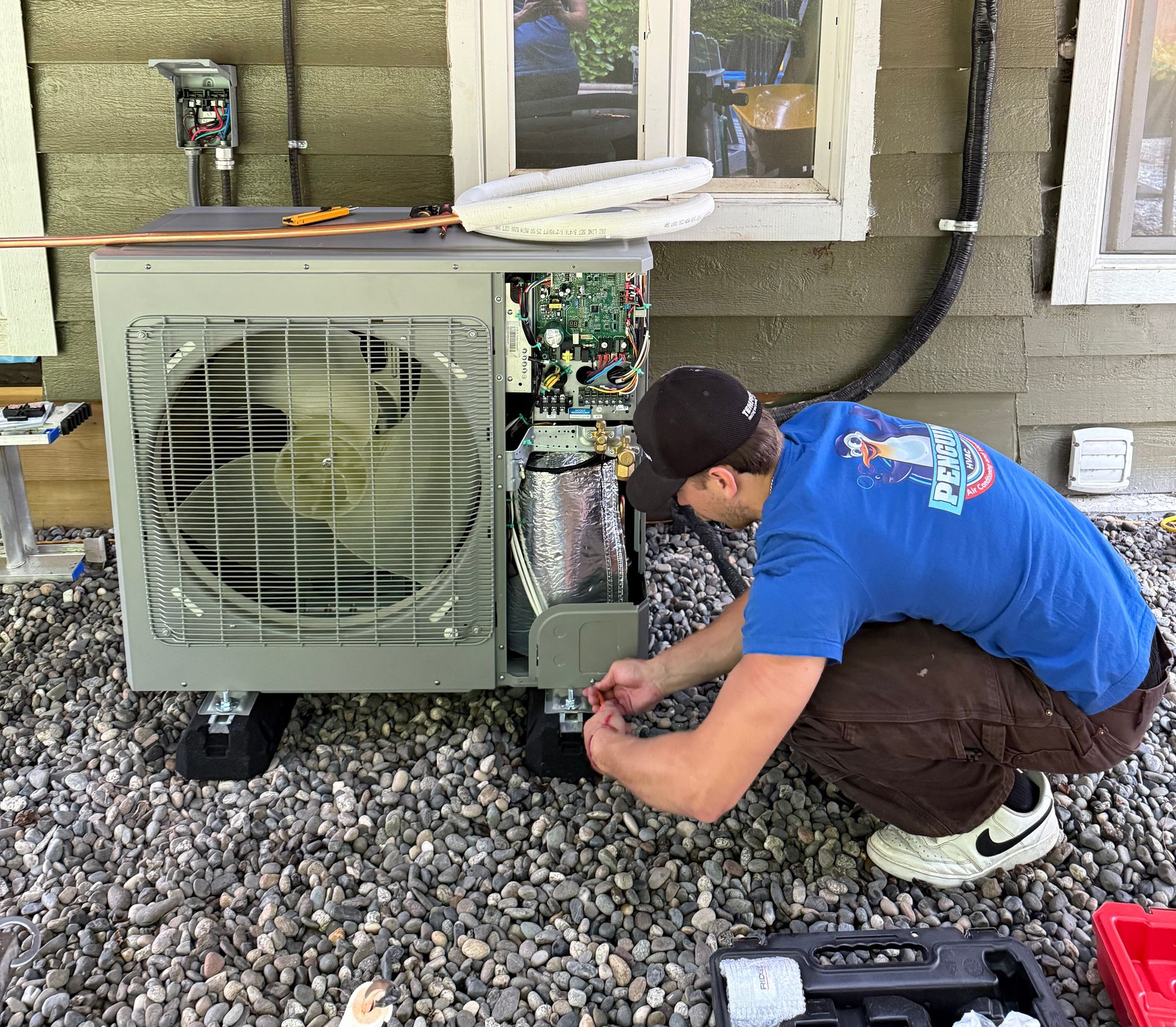 Pneguin HVAC employee installing an outdoor HVAC unit on gravel. He is kneeling, working on the exposed interior in Vancouver