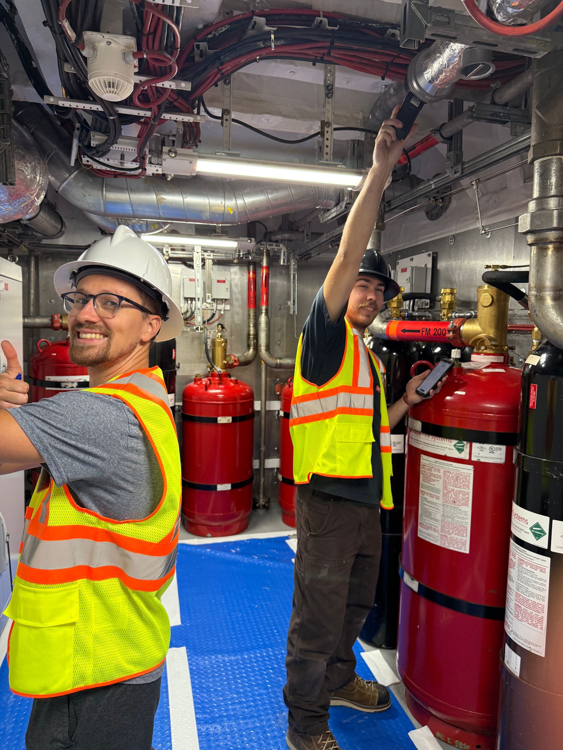 Two workers in safety vests and hard hats inspect equipment in a utility room.