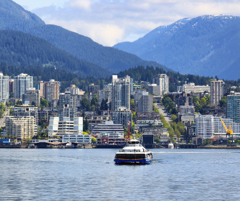Ferry boat on water in front of a city skyline and mountains under a blue sky.