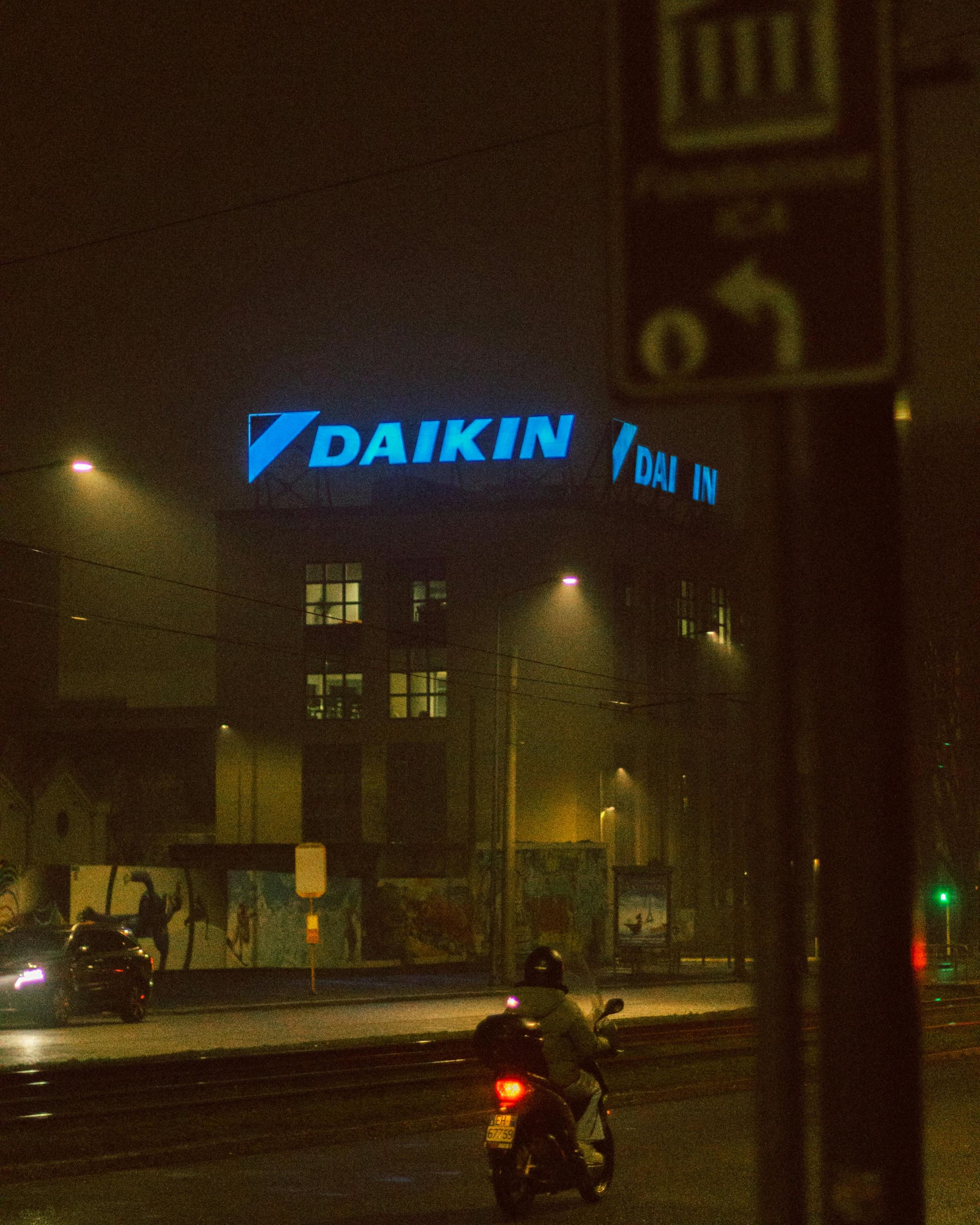 Night scene: Illuminated Daikin building sign over a wet street. A motorcycle passes, rain falling.
