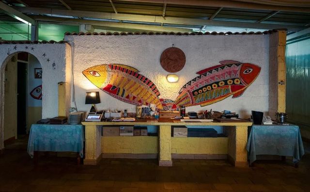 Un colorato murale raffigurante pesci si trova sopra un lungo bancone all'interno di un ristorante a tema tropicale, illuminato da luci soffuse.